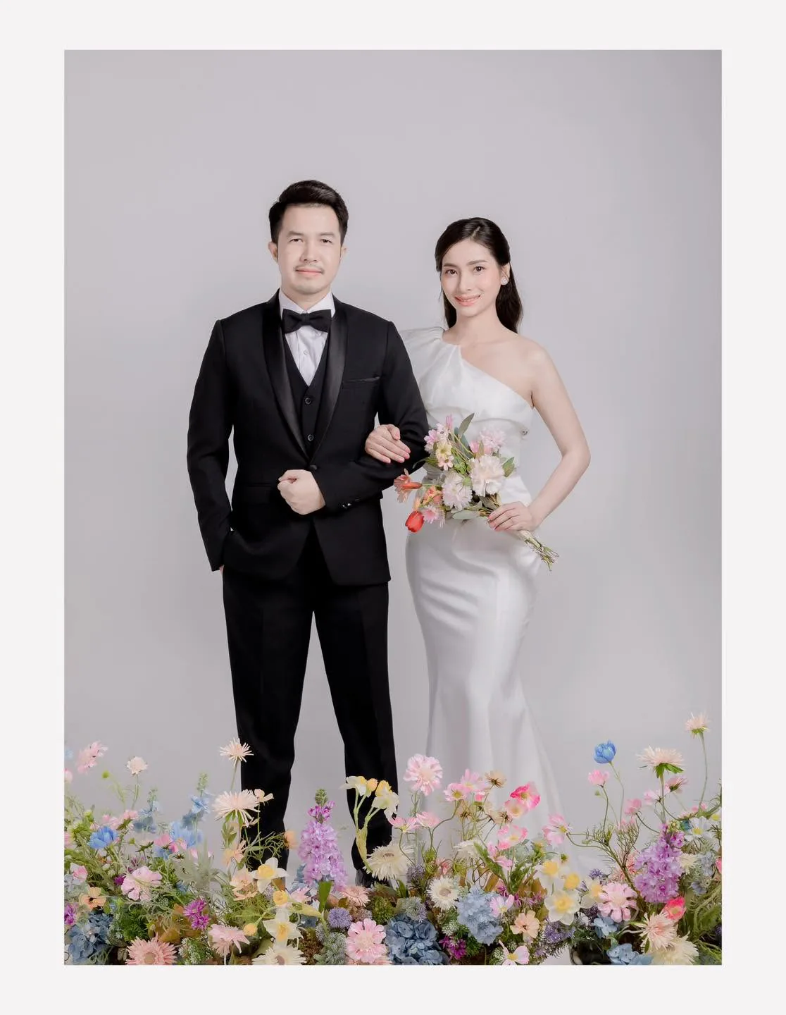 คู่บ่าวสาวยืนในชุดแต่งงานและสูท มีดอกไม้หลากสีอยู่ด้านหน้า