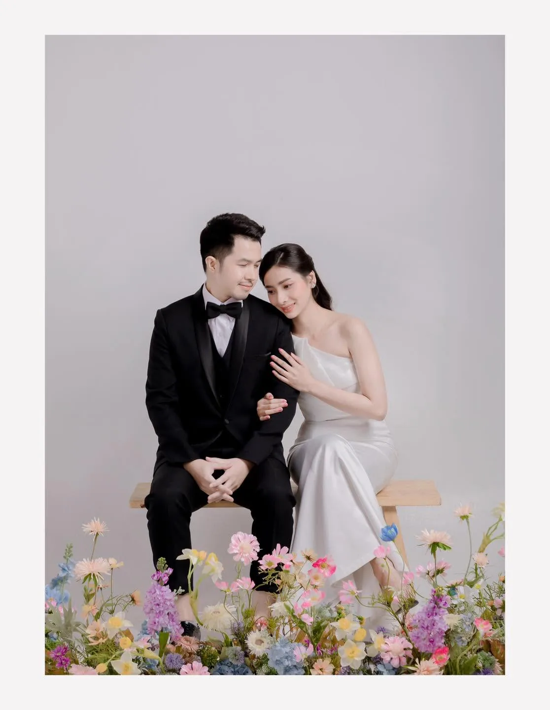 คู่บ่าวสาวนั่งพิงกันบนม้านั่งหลังแนวดอกไม้ในสตูดิโอ
