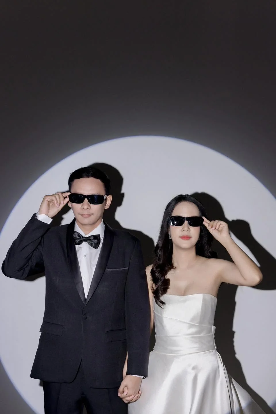 คู่บ่าวสาวสวมแว่นกันแดดในชุดแต่งงานและสูท ยืนหน้าแสงวงกลมในสตูดิโอ