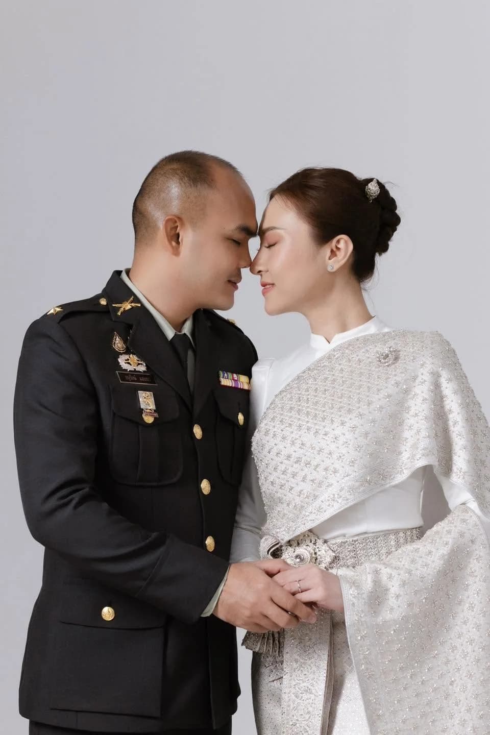 เจ้าบ่าวในเครื่องแบบทหารจับมือเจ้าสาวที่สวมชุดไทยสีเงิน
