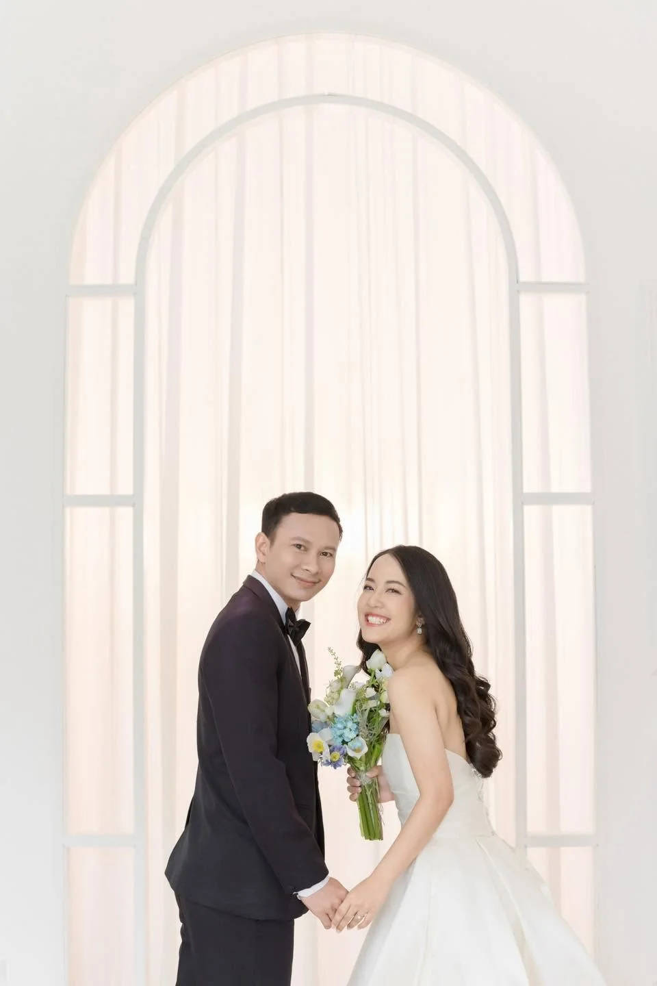 คู่บ่าวสาวยืนใกล้กันใต้ซุ้มโค้งสีขาว เจ้าสาวถือช่อดอกไม้