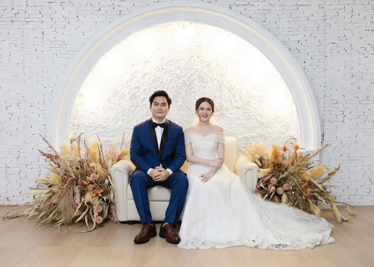 คู่บ่าวสาวนั่งคู่กันในชุดแต่งงานและสูทสีน้ำเงินหน้าซุ้มดอกไม้