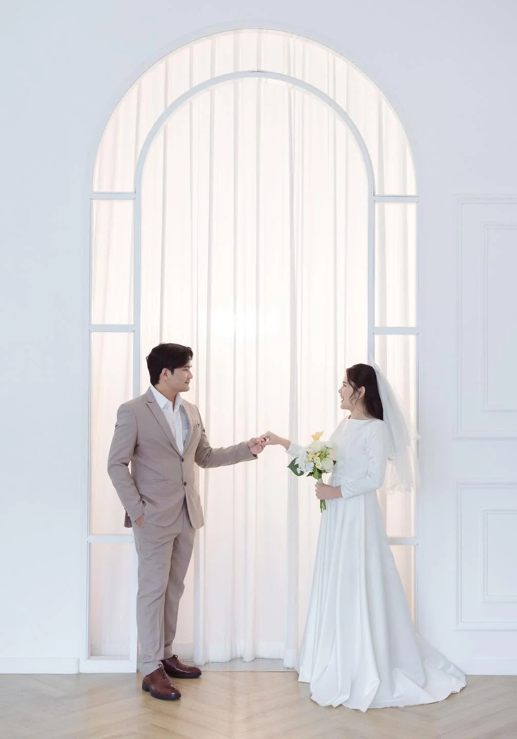 คู่บ่าวสาวยืนจับมือกันใต้ซุ้มโค้งสีขาวในสตูดิโอ